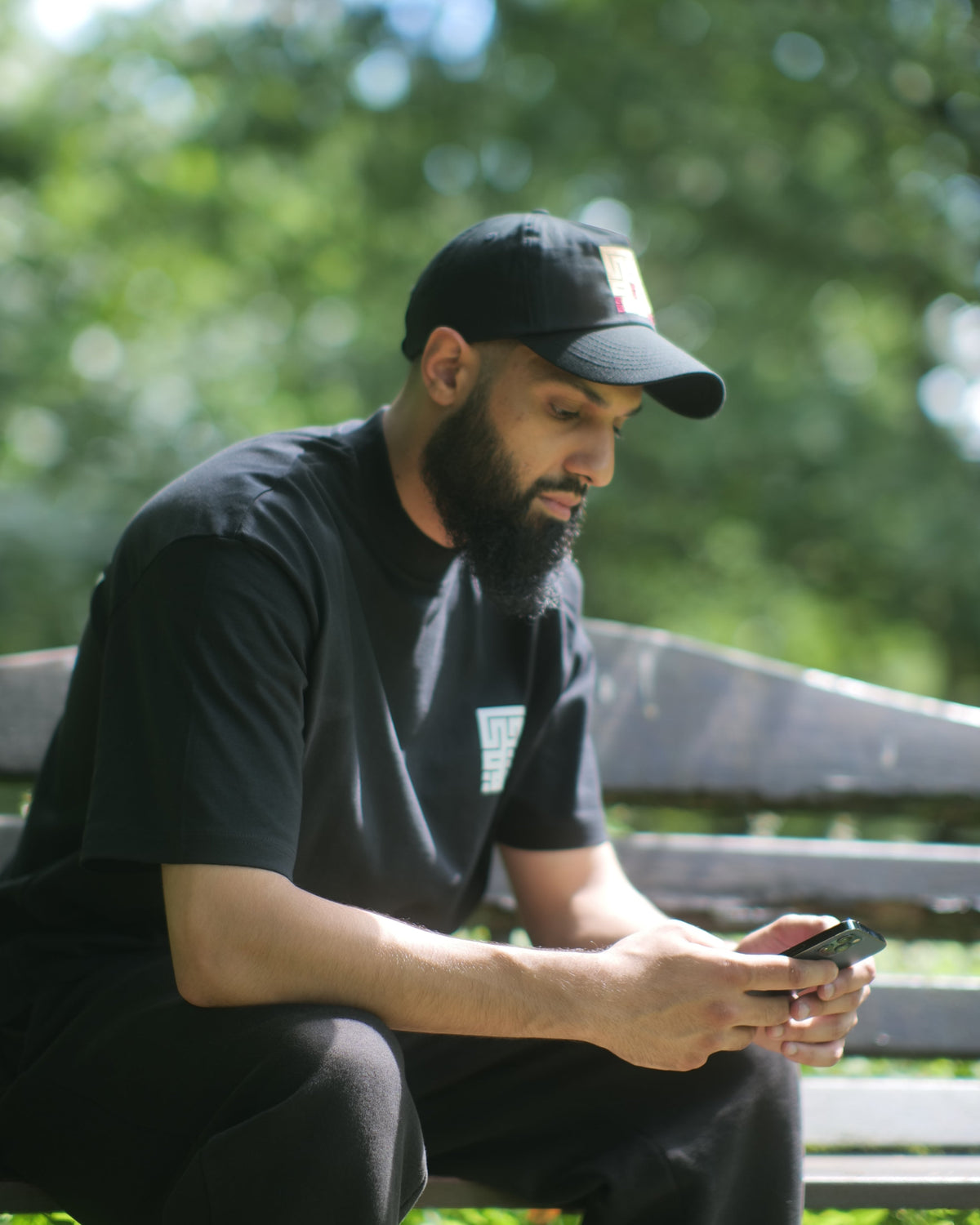 A man with a beard wearing a black t-shirt, black pants, and a black baseball cap sits on a wooden park bench, looking down at a smartphone in an outdoor setting with green foliage in the background.