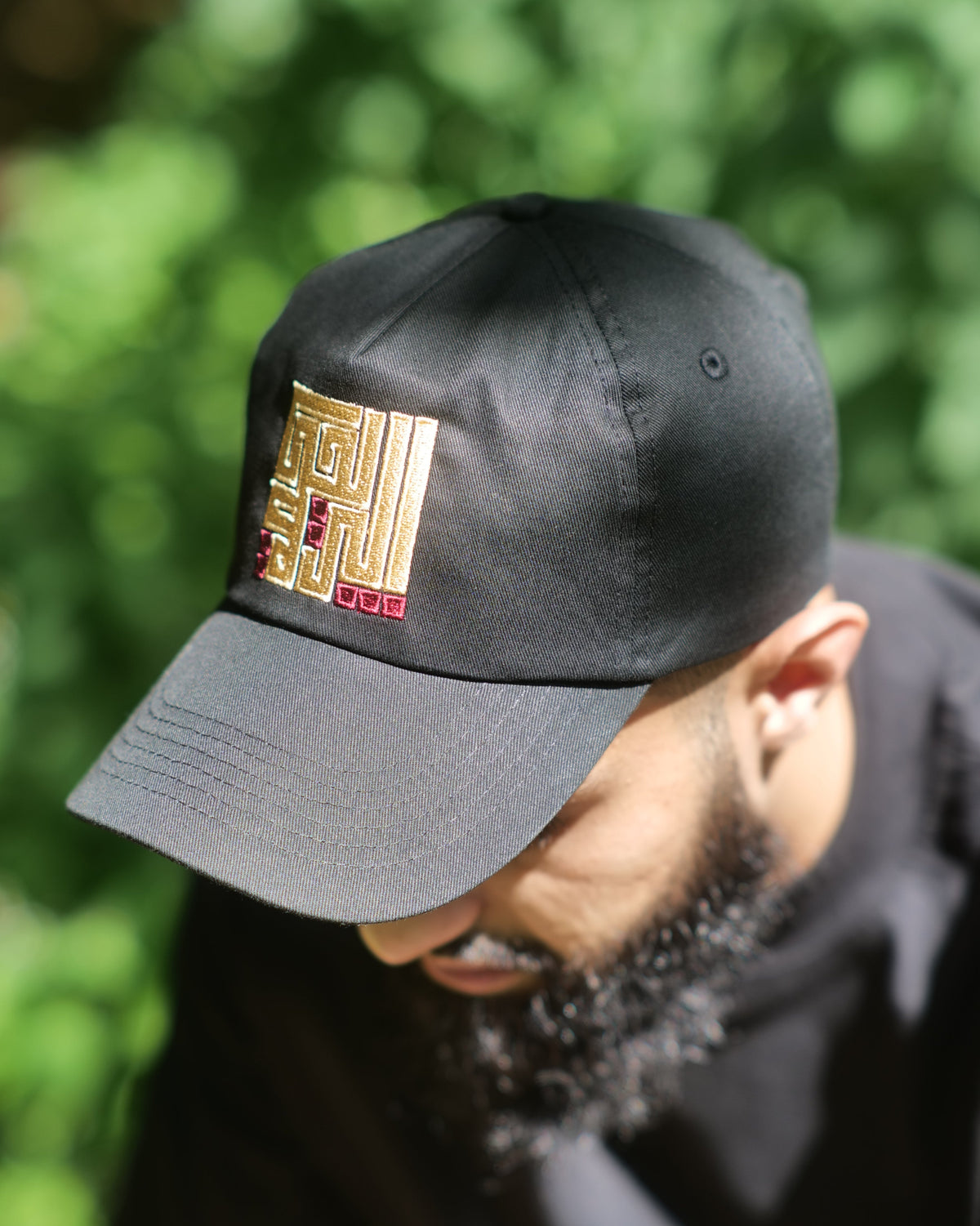 A man wearing a black baseball cap with a gold and red embroidered geometric logo on the front, photographed outdoors against a blurred green foliage background.