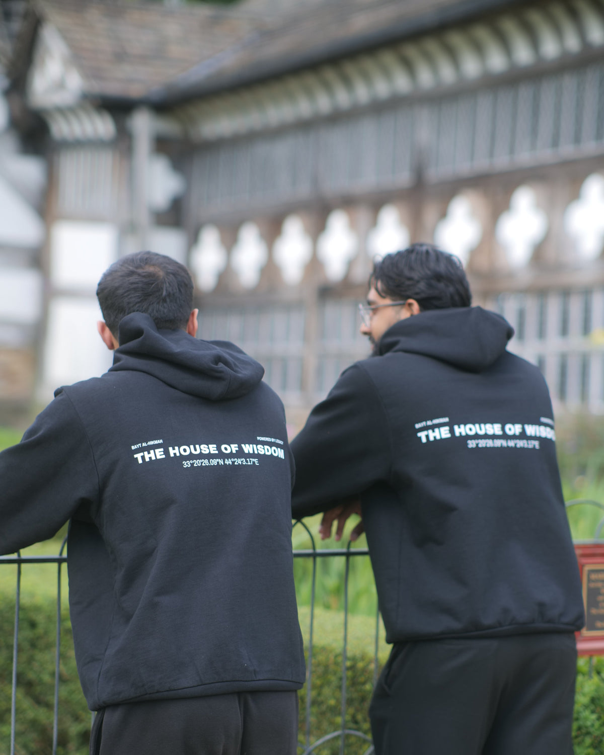 Two men wearing matching black hoodies and sweatpants stand with their backs to the camera, looking at a historic timber-framed building. The back of their clothing features white text that reads 'THE HOUSE OF WISDOM'.