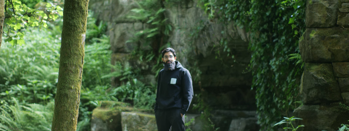 A man in a black hoodie and black pants stands on a mossy stone ruin surrounded by lush green trees and ferns, with a large stone building visible in the background.