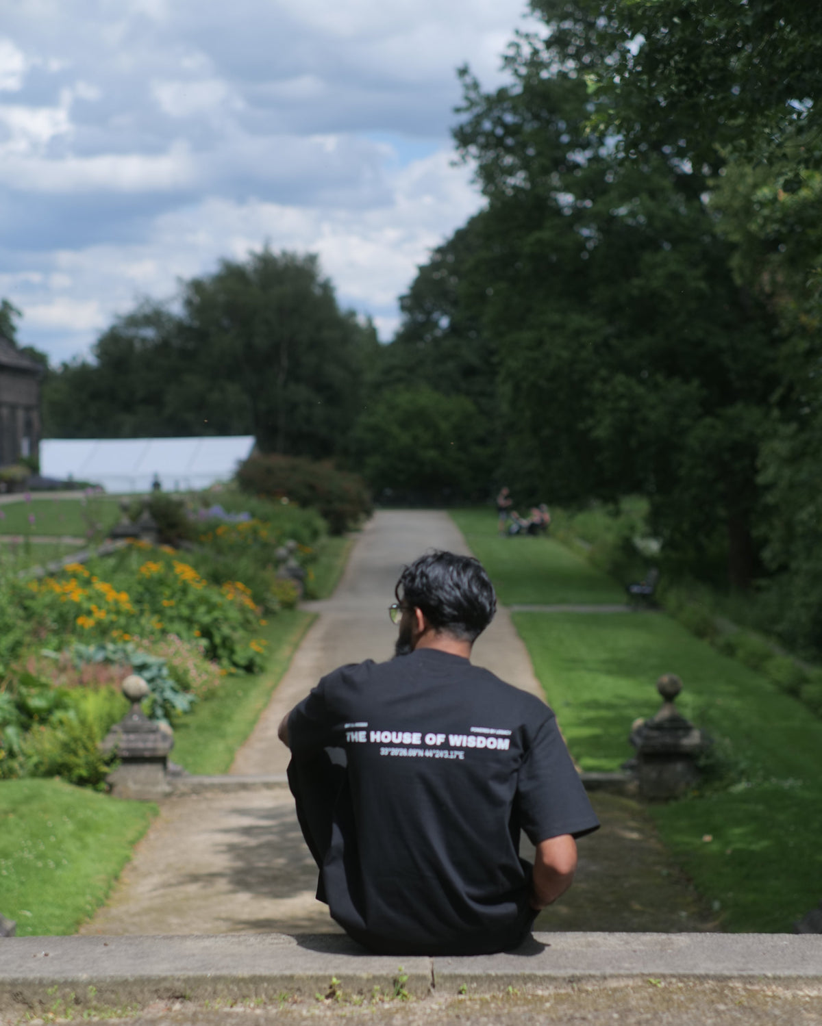 A man wearing a black t-shirt with 'HOUSE OF WISDOM' printed on the back sits on a stone wall in a garden, viewed through a circular lens or lens cover.