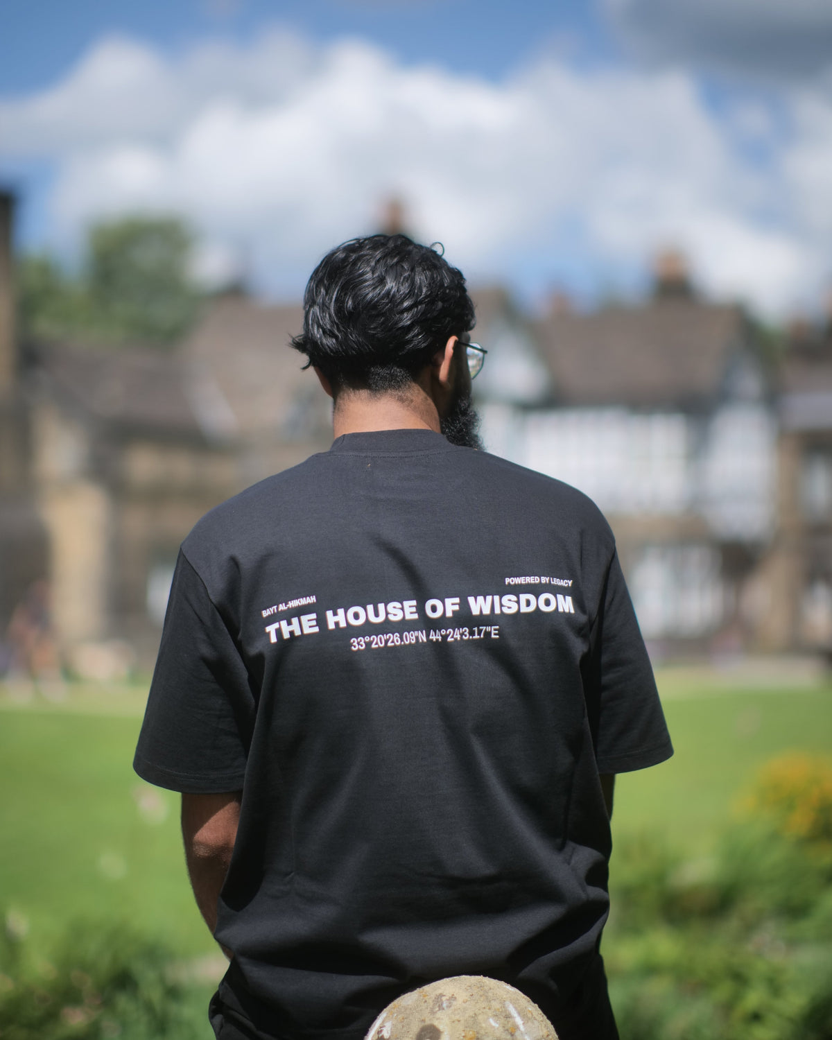 A man viewed from behind wearing a black t-shirt with 'THE HOUSE OF WISDOM' printed in white on the back, standing outdoors in a grassy park with a large stone building and blue sky in the background.