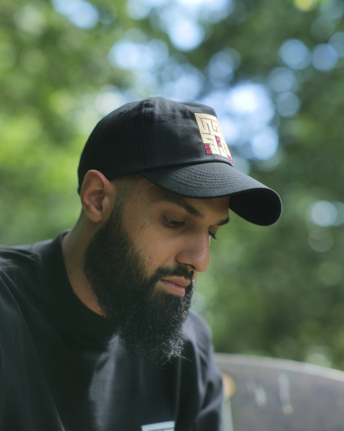 A man with a beard wearing a black baseball cap and a black t-shirt, looking down in an outdoor setting with a blurred green background.