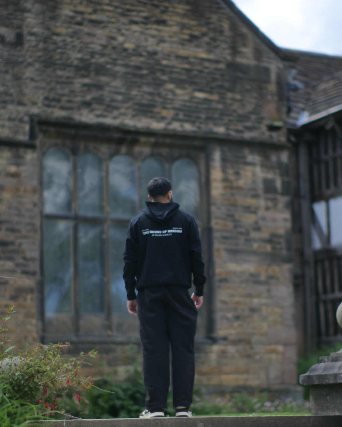 A person in a black hoodie and black pants stands on stone steps facing a large historic stone building, viewed through a circular frame with dark vignette edges.