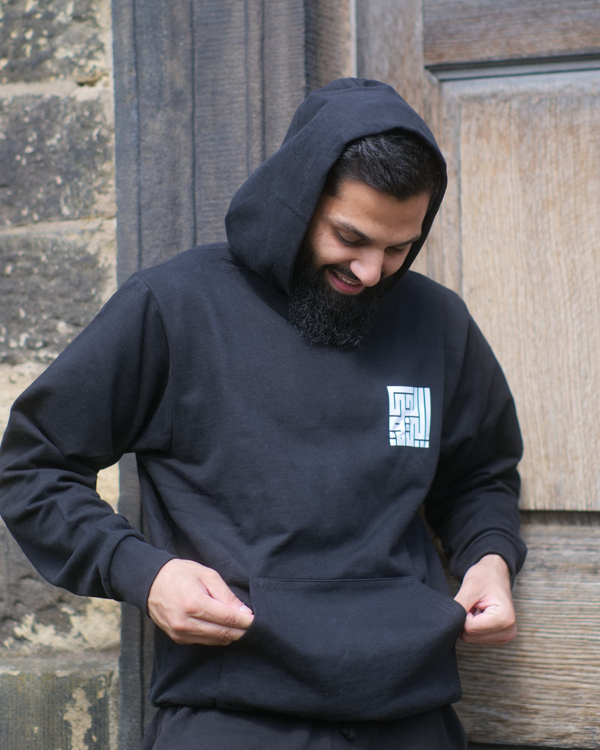 A man wearing a black hoodie and matching black sweatpants stands in front of a rustic wooden door and stone wall, pulling the hem of his hoodie down slightly. A small white square graphic is visible on the left chest.