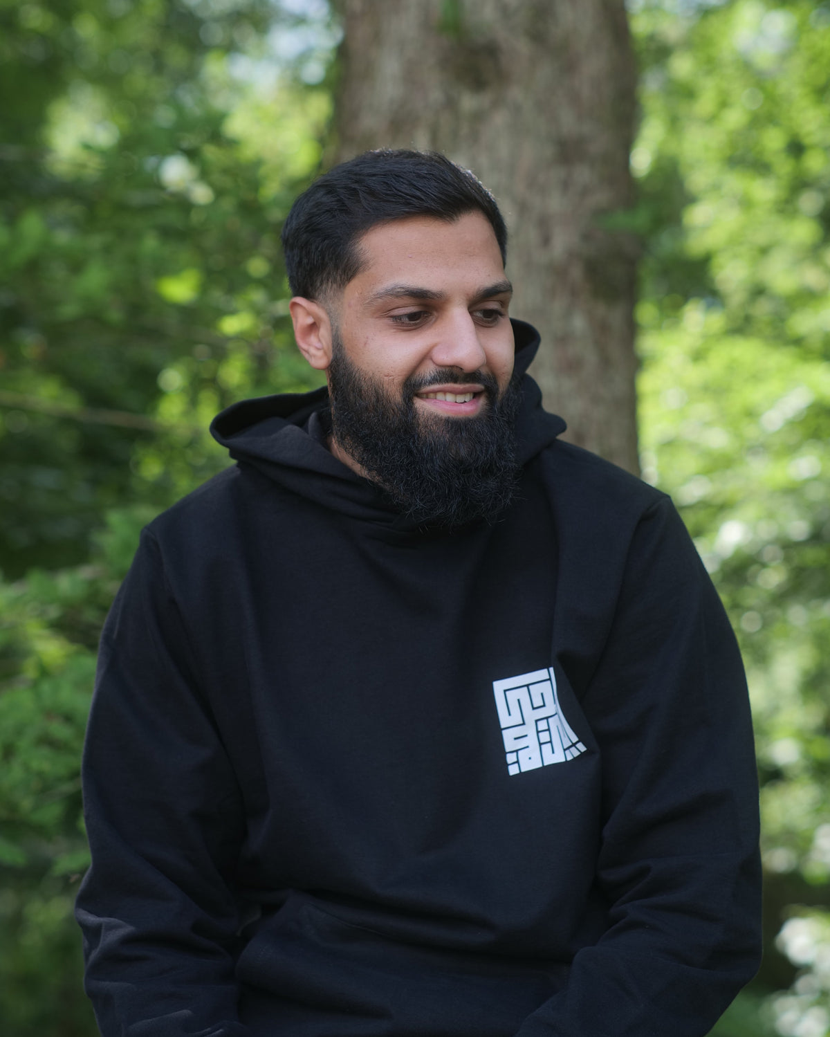 A man with a beard wearing a black hoodie with a white geometric logo on the chest, sitting outdoors in front of a blurred forest background.