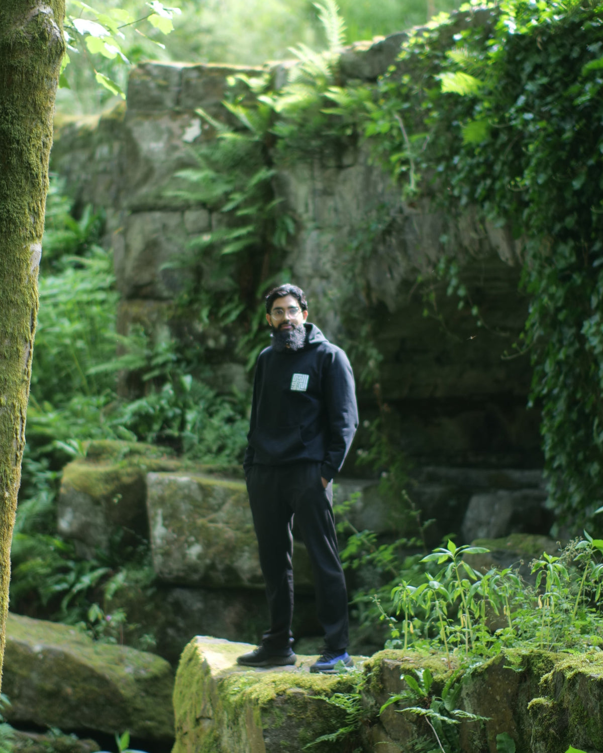 A man in a black hoodie and black pants stands on a mossy stone ruin surrounded by lush green trees and ferns, with a large stone building visible in the background.